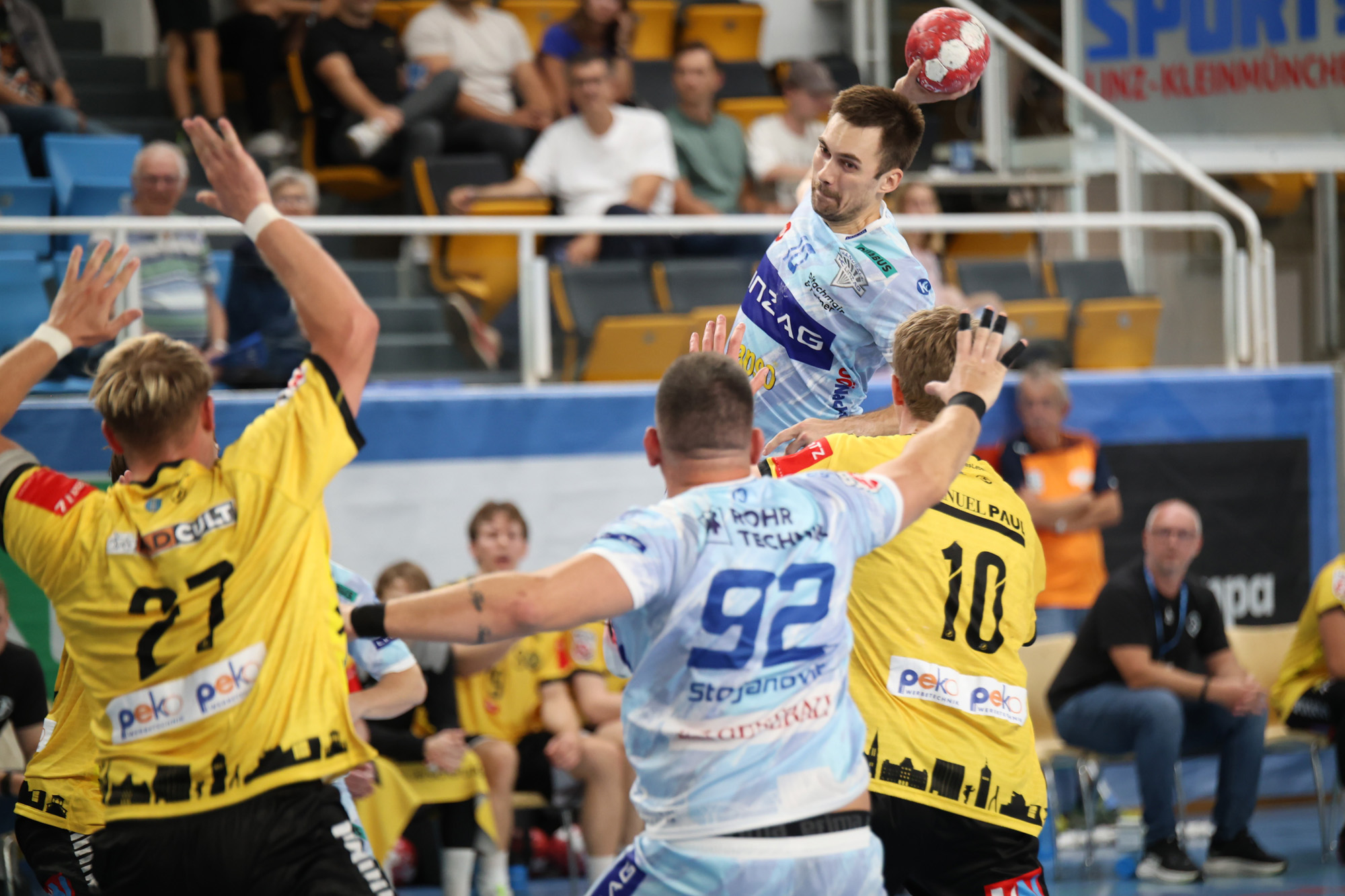Handballspieler wirft Ball, Spieler mit Rückennummer 92 und 27 im Vordergrund